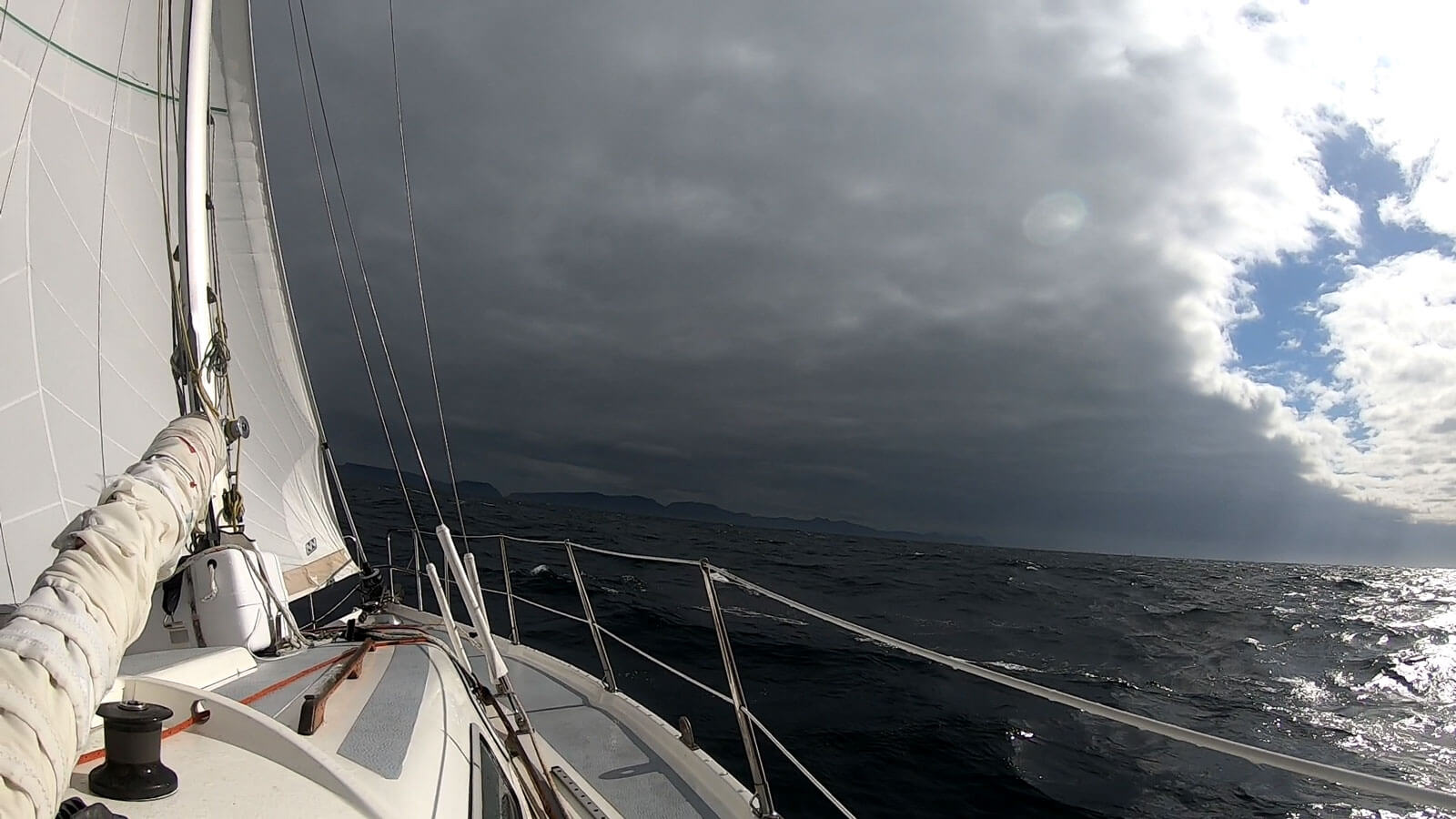Vue depuis un voilier voguant sur une mer noire, sous une couverture nuageuse importante, et des éclaircies à tribord