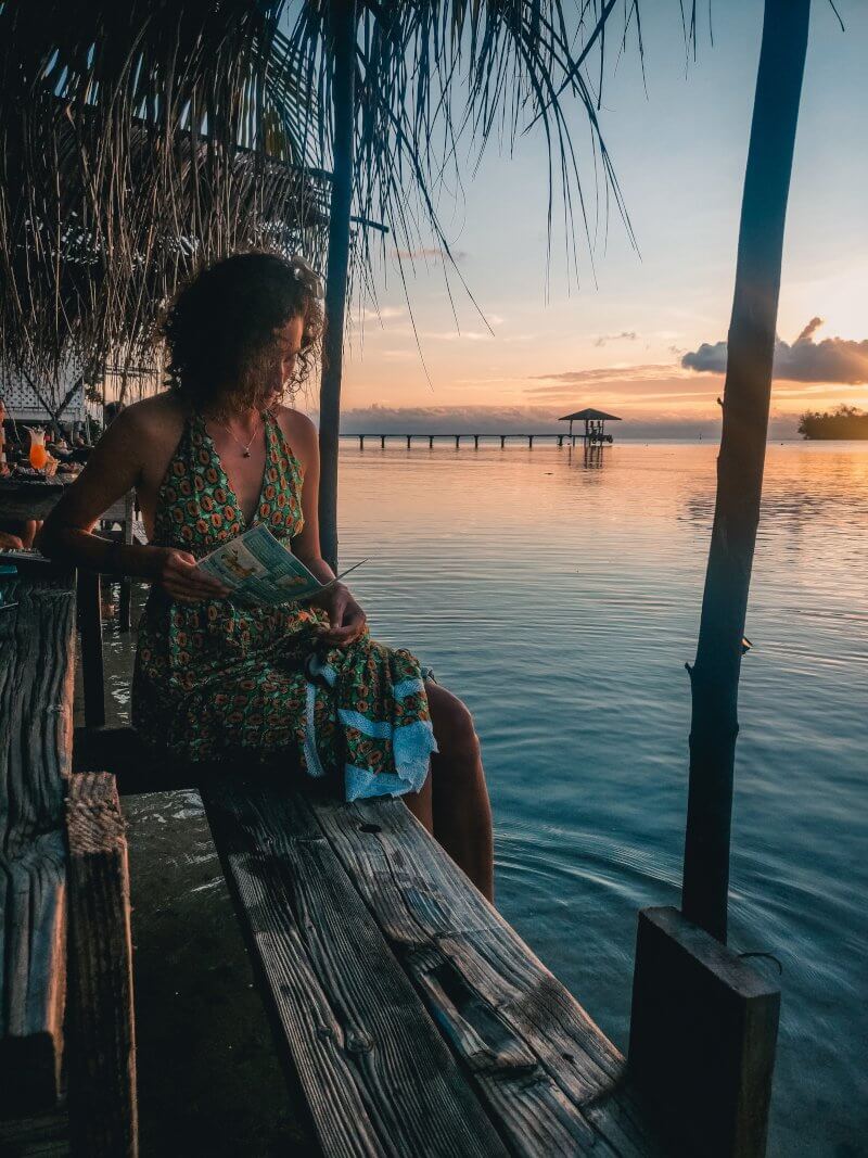 Une jeune femme lit assise sur un banc, les pieds dans l&rsquo;eau, devant le coucher de soleil