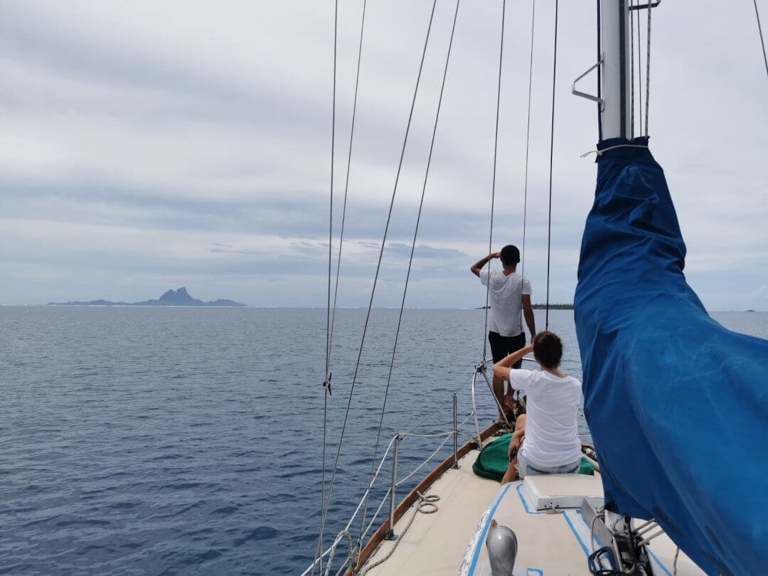 Un homme et une femme à l&rsquo;avant d&rsquo;un voilier, les mains en visière, observent l&rsquo;île de Bora-bora à l&rsquo;horizon