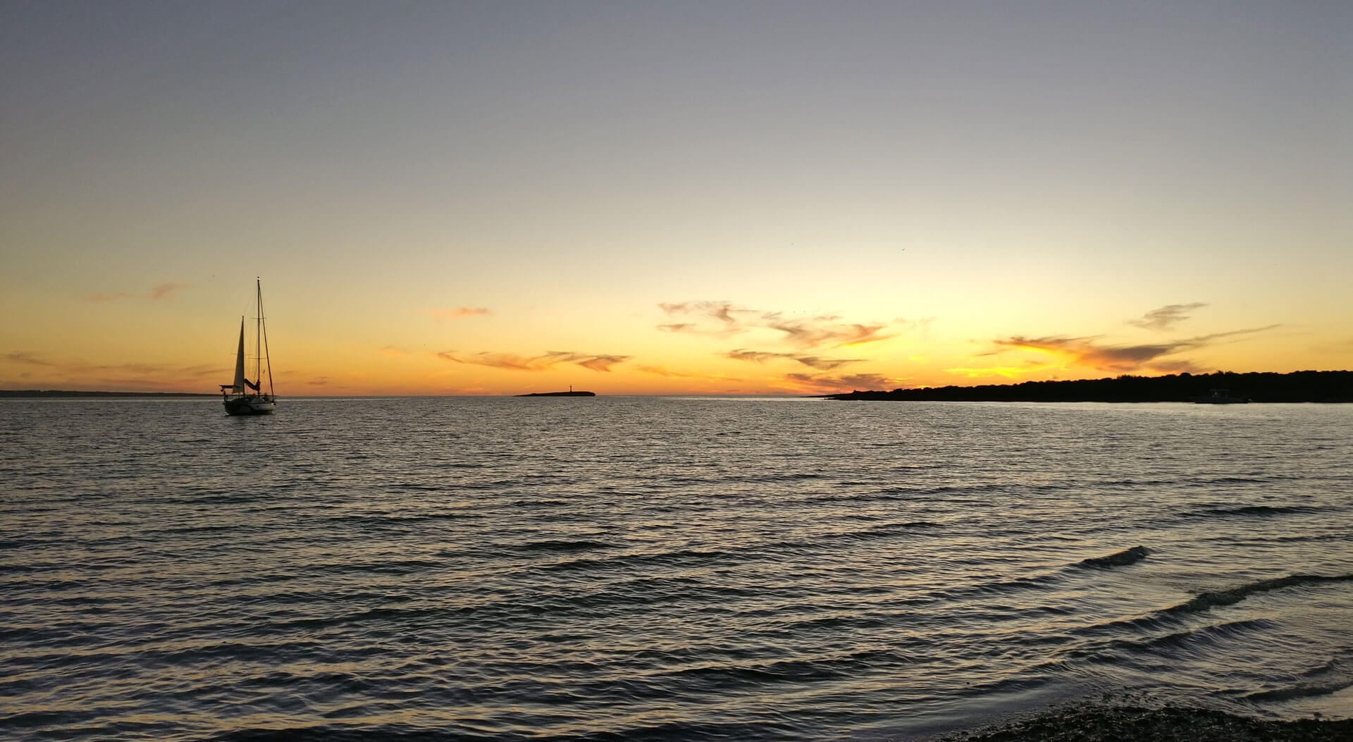 Le crépuscule baigne l&rsquo;horizon d&rsquo;une douce lumière jaune, et on voit un voilier au loin